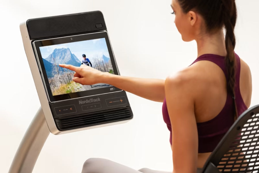 Woman in purple sports bra using NordicTrack recumbent exercise bike with touchscreen display showing mountain landscape, on 