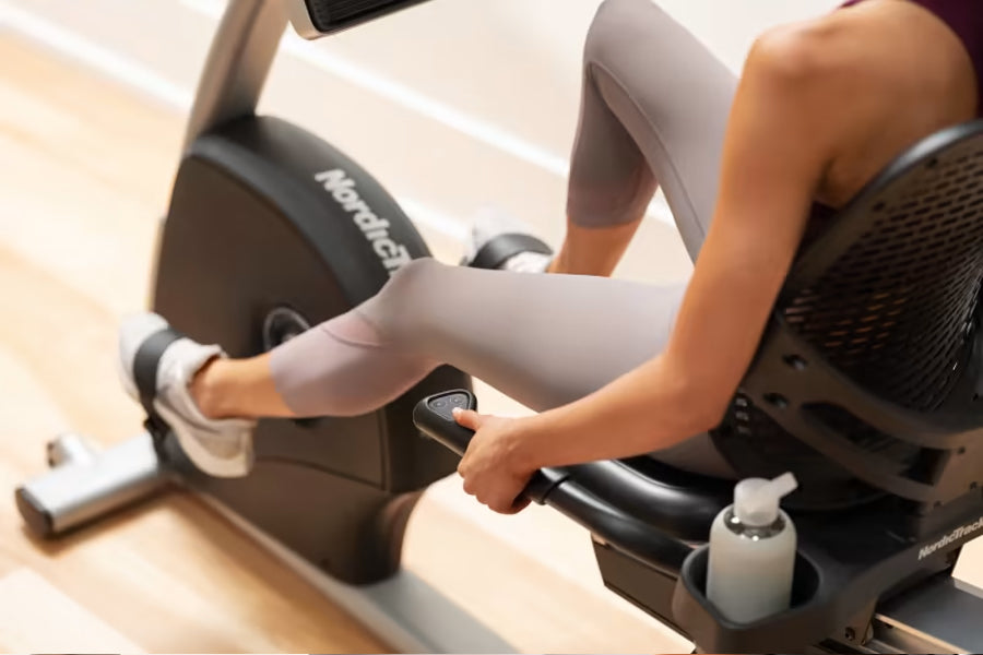 Person in white workout leggings using black NordicTrack recumbent exercise bike with hand grips and display console