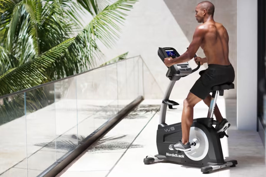 Shirtless man on black and white NordicTrack upright exercise bike with digital console, in modern room with palm plants