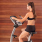 Woman in black sports bra and shorts using silver and black upright exercise bike against wooden wall background