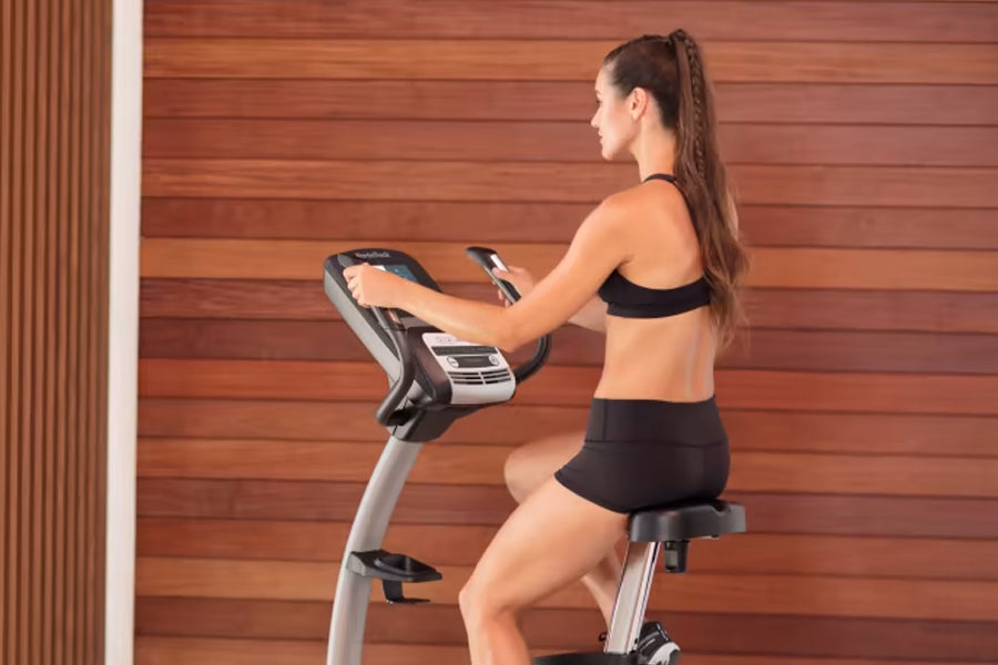 Woman in black sports bra and shorts using silver and black upright exercise bike against wooden wall background