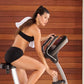 Woman wearing black sports bra and shorts with white headband riding upright exercise bike against wooden wall background