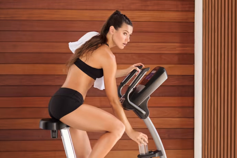 Woman wearing black sports bra and shorts with white headband riding upright exercise bike against wooden wall background