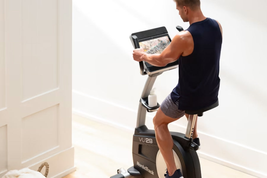 Man in navy sleeveless shirt using NordicTrack VU 29 upright exercise bike with digital display screen in home gym setting