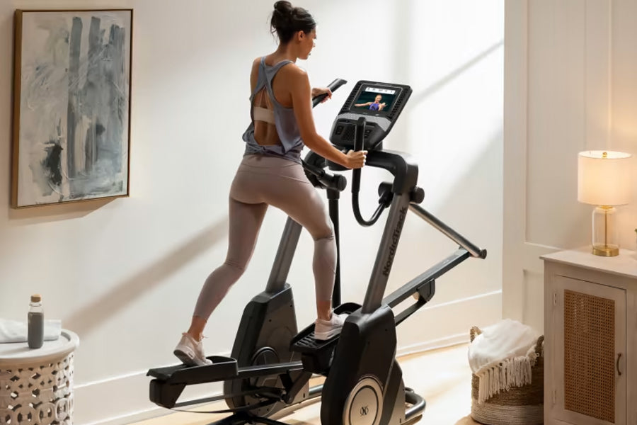 Woman wearing gray sports bra and leggings using black NordicTrack elliptical machine with digital display in bright home gym