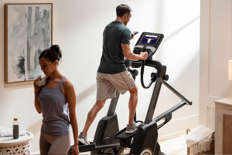 Man on black elliptical machine with digital display in home gym, woman in gray tank top in foreground
