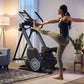 Woman using black NordicTrack elliptical machine with extended arms in modern living room setting