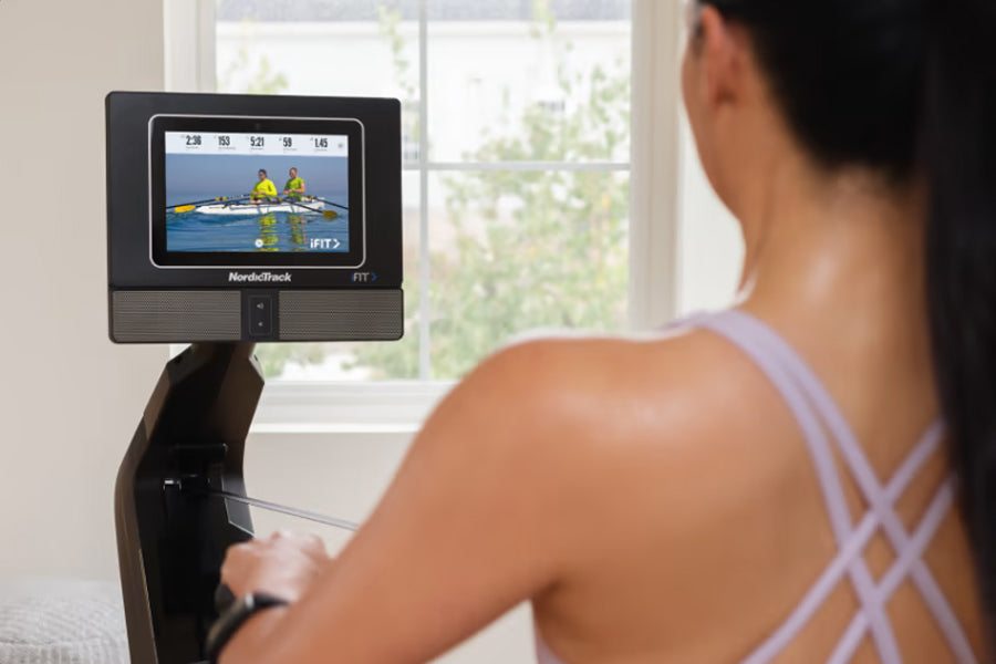 Woman in lavender sports bra using NordicTrack rowing machine with display screen showing rowing workout video