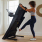 Black motorized treadmill with digital display, woman in gray sports bra and navy leggings using it in bright home gym