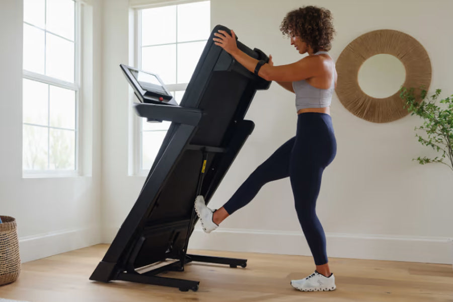 Black motorized treadmill with digital display, woman in gray sports bra and navy leggings using it in bright home gym