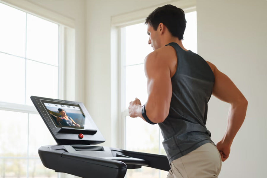 Man wearing black sleeveless tank top running on treadmill with digital display in bright room with windows
