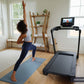Black motorized treadmill with digital display, shown in home gym setting with woman exercising on blue mat
