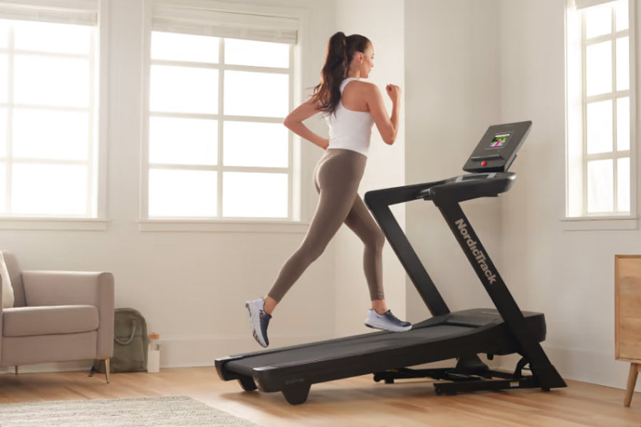 Woman running on black NordicTrack treadmill wearing white sports bra and beige leggings in bright home gym