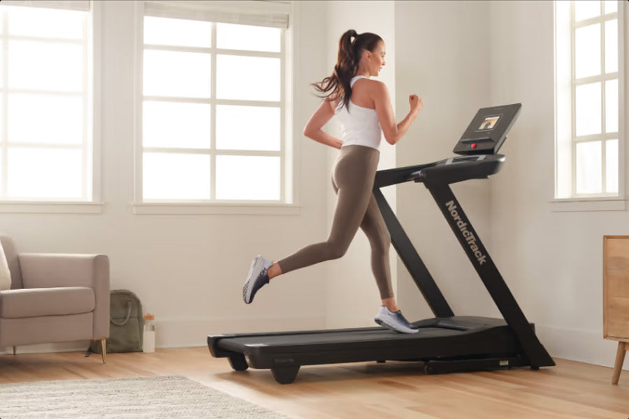 Woman running on black NordicTrack treadmill wearing white sports bra and brown leggings in bright home gym