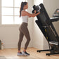 Woman wearing white tank top and brown leggings using black NordicTrack EXP 7i treadmill with dumbbells in bright home gym