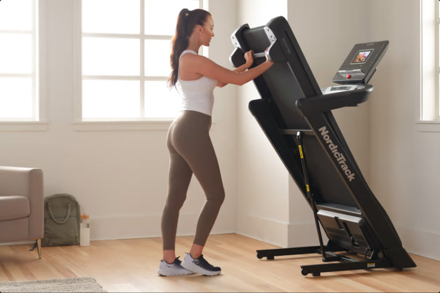 Woman wearing white tank top and brown leggings using black NordicTrack EXP 7i treadmill with dumbbells in bright home gym