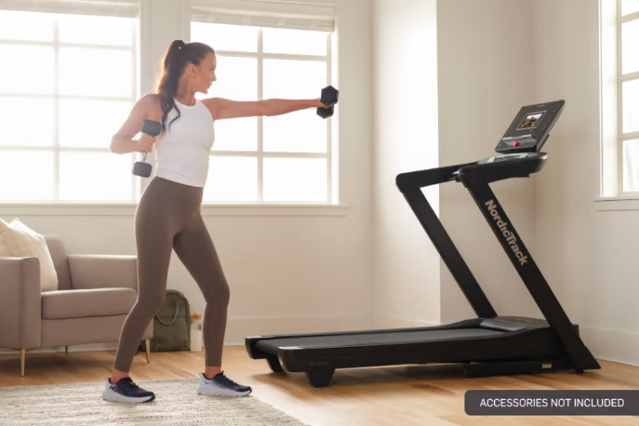 Woman wearing white tank top and brown leggings exercising with dumbbells in front of black NordicTrack treadmill in home gym