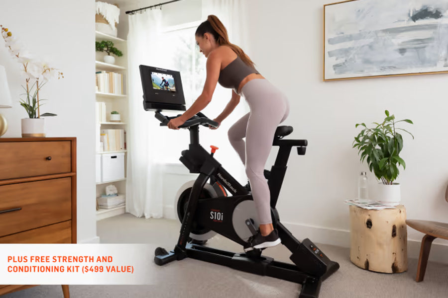 Woman wearing brown sports bra and light gray leggings riding black NordicTrack S10i stationary bike with digital display in 