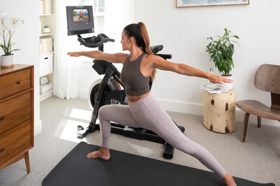 Woman performing lunge exercise on black mat in front of NordicTrack S10i stationary bike with screen display in home gym set