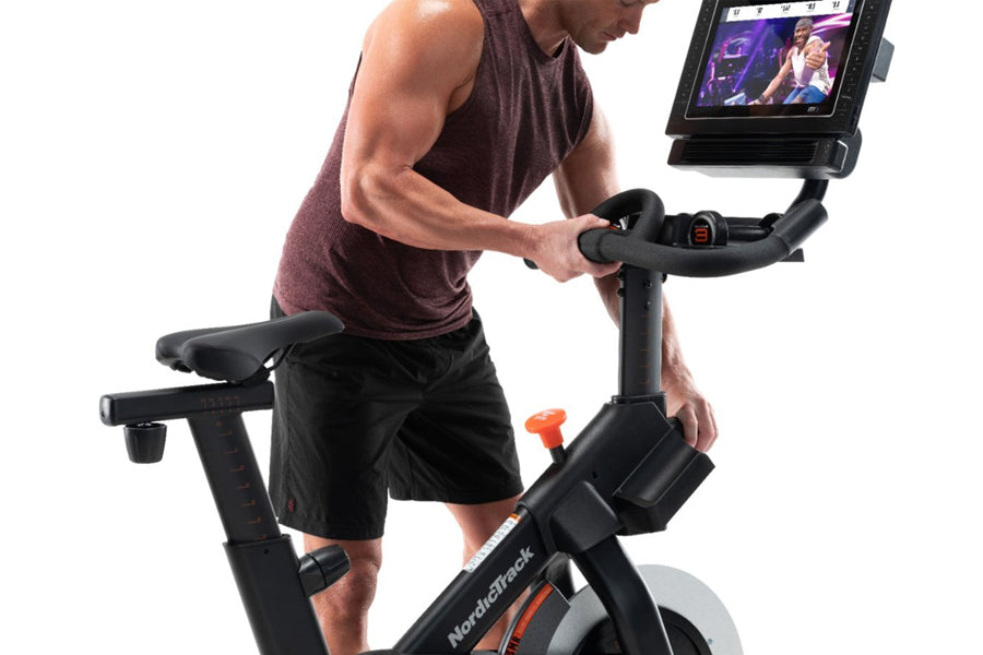Man in burgundy tank top and black shorts using NordicTrack S15i stationary bike with tablet display showing workout content