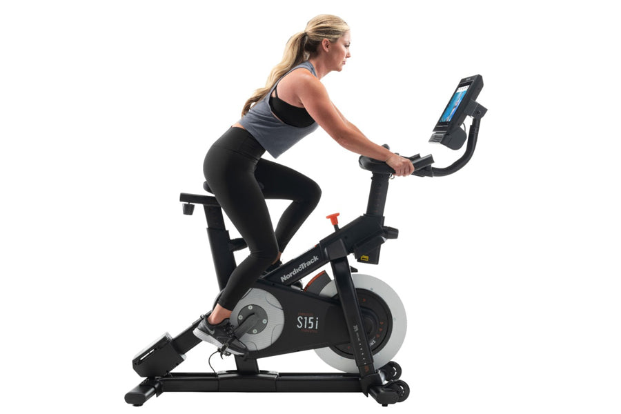 Woman on black NordicTrack S15i stationary bike with digital display, wearing black sports bra and leggings, on white backgro