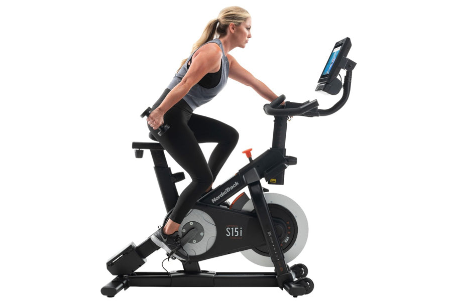 Woman using black NordicTrack S15i stationary exercise bike with digital display, worn by model on white background