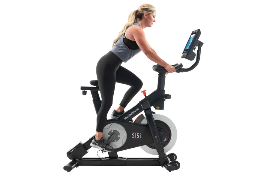 Woman in black leggings and gray sports bra riding NordicTrack S15i stationary exercise bike with blue digital display on whi