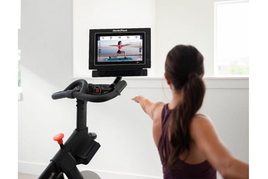 Black NordicTrack stationary exercise bike with digital screen displaying fitness class, shown with woman using it indoors