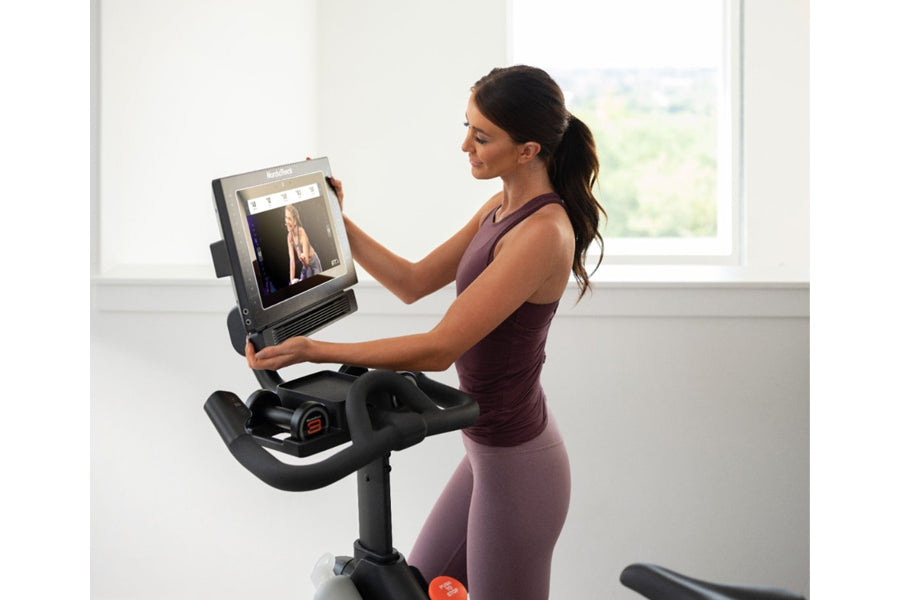 Woman in purple athletic wear using NordicTrack S15i stationary bike with touchscreen display showing workout class