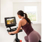 Woman using stationary exercise bike with digital display screen showing outdoor cycling video, wearing burgundy workout tank