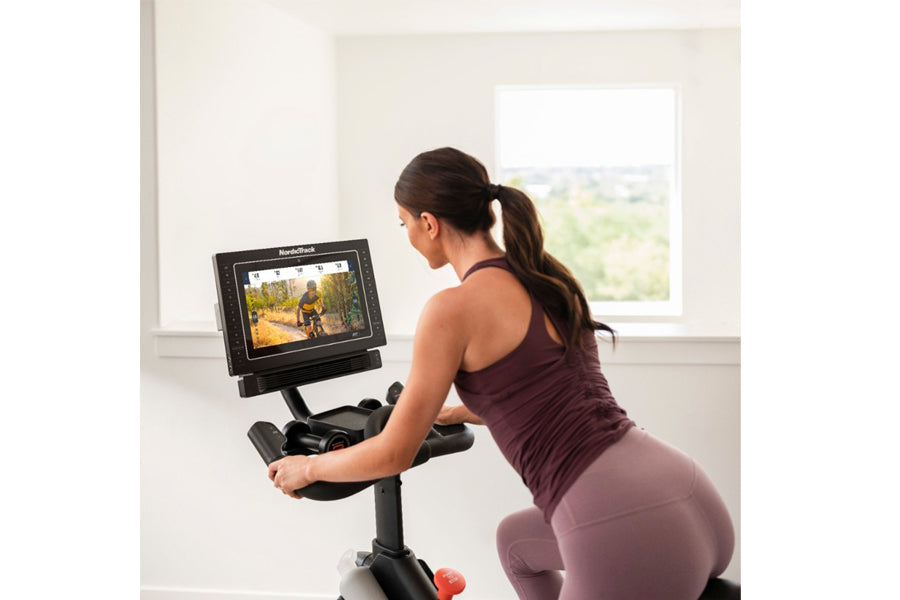 Woman using stationary exercise bike with digital display screen showing outdoor cycling video, wearing burgundy workout tank