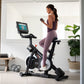 Woman in purple athletic wear exercising on black NordicTrack S15i stationary bike with tablet display, holding dumbbell in b