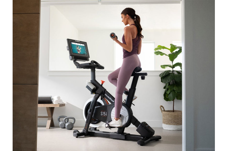 Woman in purple athletic wear exercising on black NordicTrack S15i stationary bike with tablet display, holding dumbbell in b