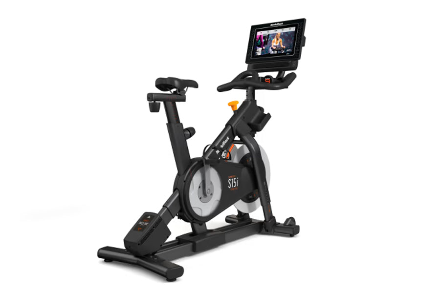 Black NordicTrack S15i indoor exercise bike with attached touchscreen display showing workout content, viewed from side on wh