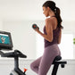 Woman wearing purple tank top and leggings on stationary exercise bike holding dumbbell, screen displaying workout video