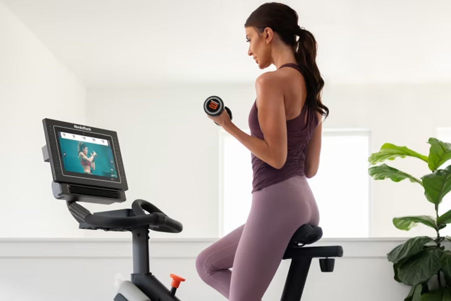 Woman wearing purple tank top and leggings on stationary exercise bike holding dumbbell, screen displaying workout video
