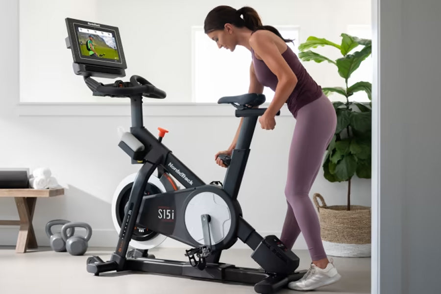 Woman on black NordicTrack S15i stationary bike with attached tablet display, wearing purple leggings and tank top in home gy
