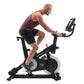 Man wearing burgundy tank top and black shorts using black NordicTrack S15i stationary bike with digital display on white bac