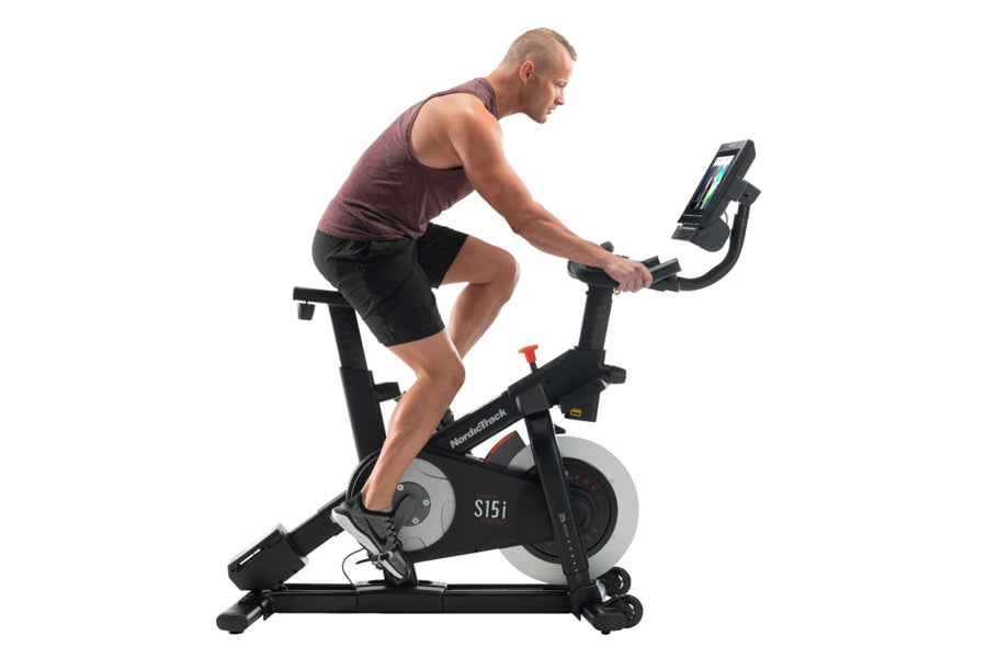 Man wearing burgundy tank top and black shorts using black NordicTrack S15i stationary bike with digital display on white bac