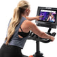 Woman using NordicTrack S15i stationary bike with touchscreen display showing workout content, wearing gray tank top and blac