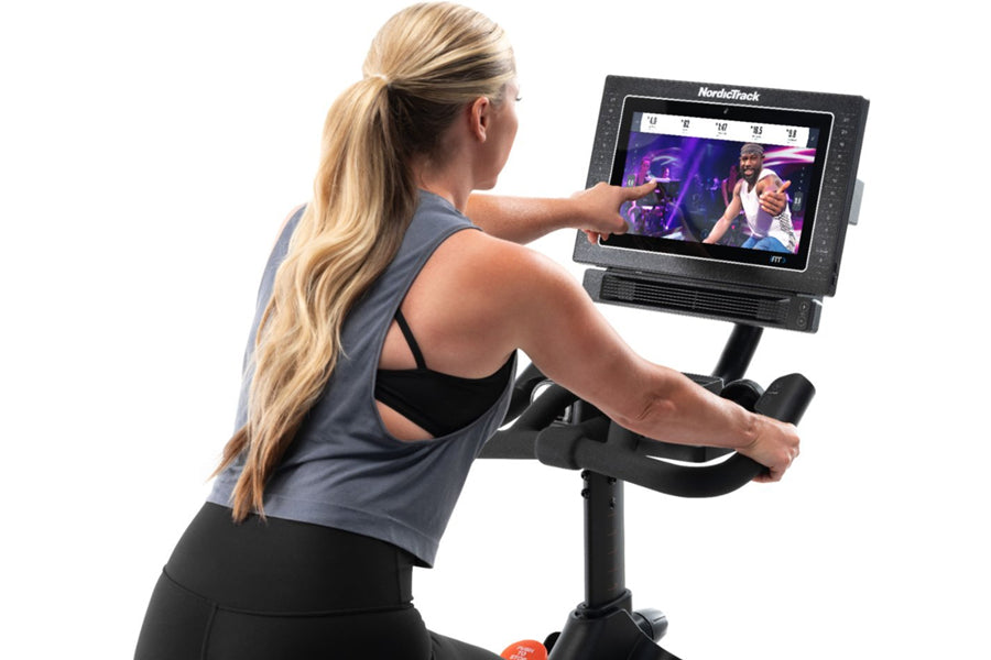 Woman using NordicTrack S15i stationary bike with touchscreen display showing workout content, wearing gray tank top and blac