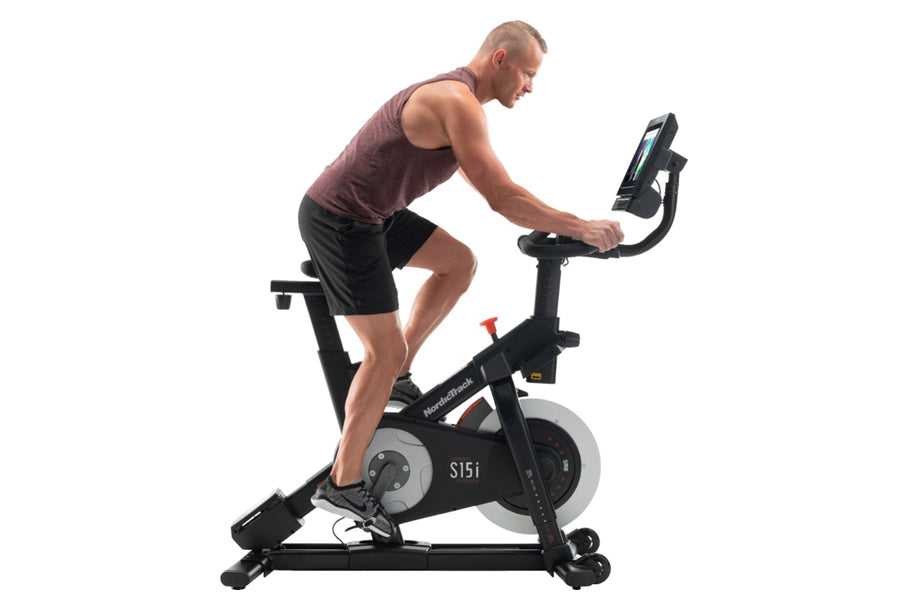 Man in burgundy tank top and black shorts riding black NordicTrack S15i stationary bike with digital display on white backgro