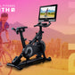 Black stationary exercise bike with digital touchscreen display showing outdoor cycling video, displayed on gradient pink to 
