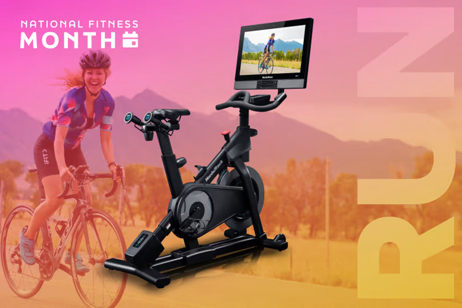 Black stationary exercise bike with digital touchscreen display showing outdoor cycling video, displayed on gradient pink to 