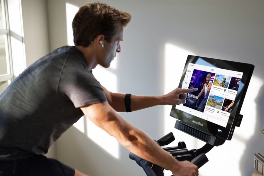 Man wearing gray t-shirt and black armband on stationary bike with touchscreen display showing streaming content