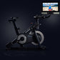 Black stationary exercise bike with grey flywheel and touchscreen display mounted above, shown on dark background