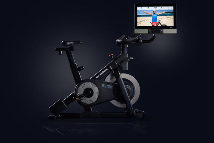 Black stationary exercise bike with grey flywheel and touchscreen display mounted above, shown on dark background
