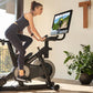 Woman in black athletic wear riding NordicTrack S27i stationary bike with tablet display, in home gym with windows and plants