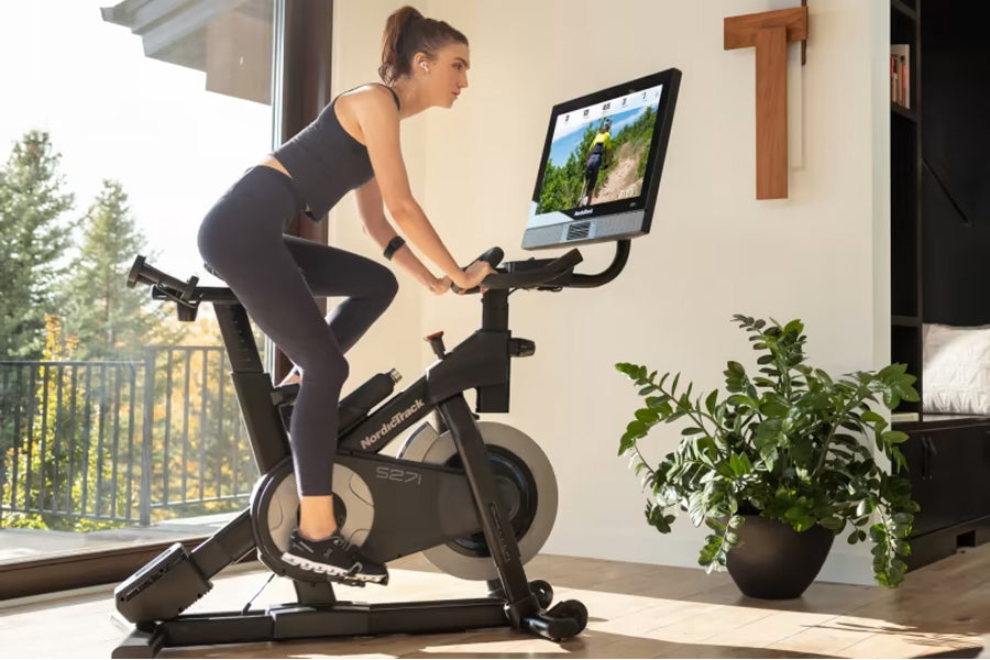 Woman in black athletic wear riding NordicTrack S27i stationary bike with tablet display, in home gym with windows and plants
