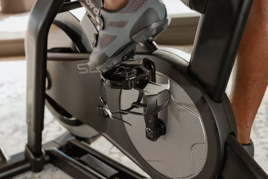 Black and gray stationary exercise bike with digital display and pedals, shown with person's feet in gray sneakers positioned